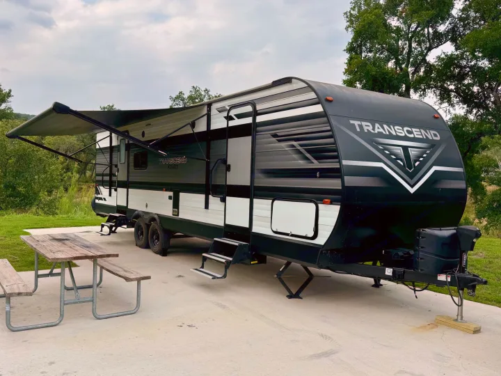 This RV rental, a black and white Transcend travel trailer, is parked at a scenic campsite with an awning and picnic table.. Grand Design Transcend Xplor 2022