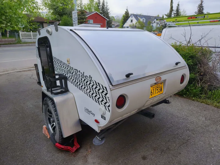 A white LittleGuy Rough Rider teardrop camper, perfect for an RV rental, with an Alaska license plate. Secured by a tire claw.. Little Guy Other 2012