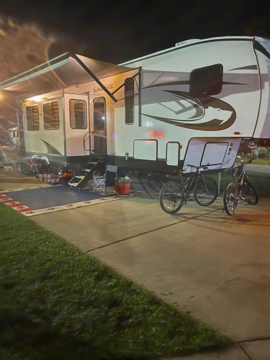 Nighttime scene of a large RV rental parked at a campsite, featuring an illuminated awning, outdoor rug, and two bikes.. Forest River Sabre 2024