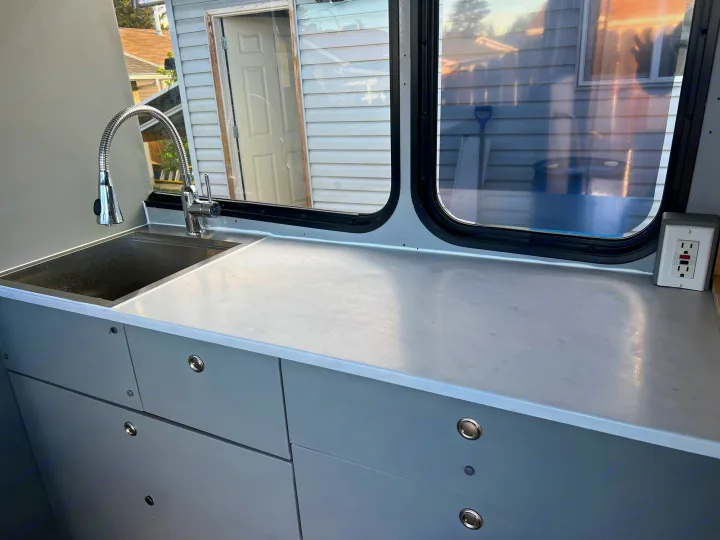 RV rental's clean interior: Light gray kitchen with sink, faucet, countertop, cabinets, and windows showing outdoor scene.. Chevrolet Express shuttle 2014