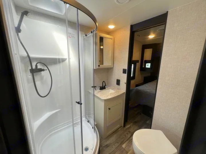 RV rental bathroom with a compact curved glass shower, vanity sink, and toilet. A bedroom is visible through the doorway.. Open Range Lite 2023