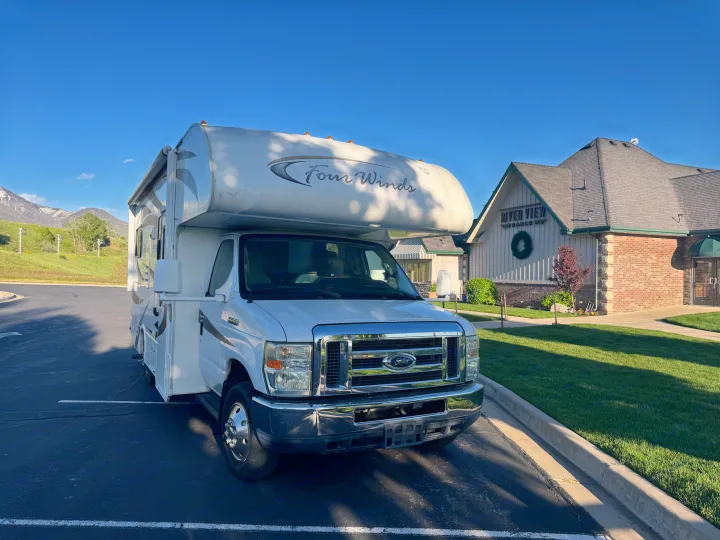 Class C Four Winds RV rental parked in front of a River View building with scenic mountains behind.. Thor Four Winds 2014