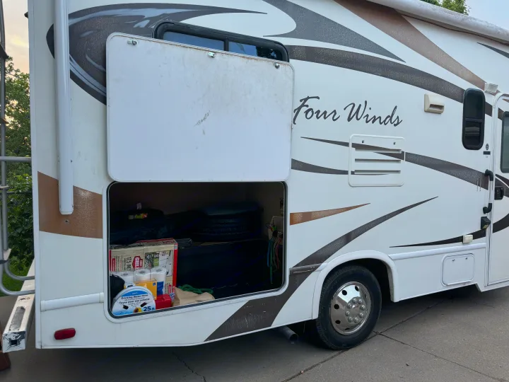 White Four Winds RV rental with an open storage bay. Inside, camping essentials like a water hose and toilet paper are visible.. Thor Four Winds 2014