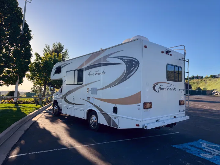 A clean, white Four Winds RV rental is parked in a sunlit lot with green trees & blue sky.. Thor Four Winds 2014