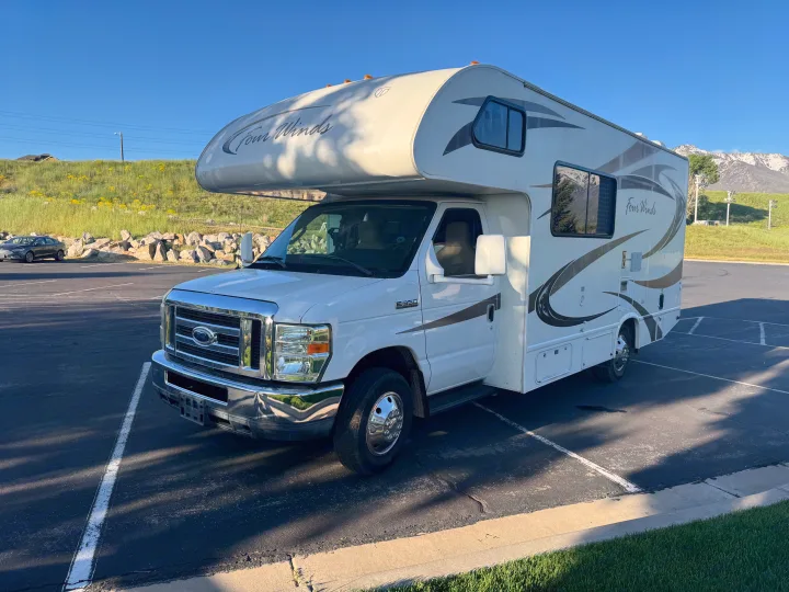 This white Four Winds Class C RV rental (Ford E350) is parked in a lot, with green hills and distant mountains under a blue sky.. Thor Four Winds 2014