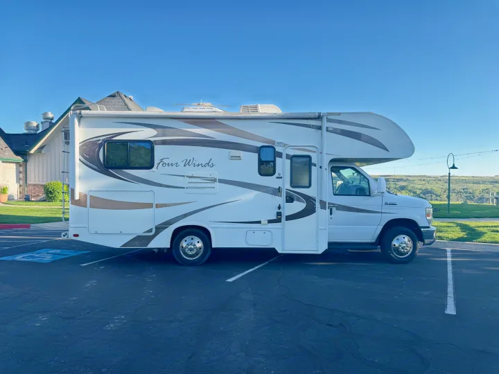 White "Four Winds" RV rental motorhome parked in a paved lot under a clear blue sky, with a building behind it.. Thor Four Winds 2014