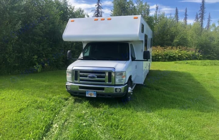 This white RV rental is parked on a lush green lawn, backed by dense trees under a blue sky, ready for outdoor travel.. Thor Majestic 2016