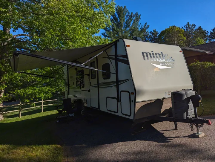 A 'Mini Lite Rockwood' travel trailer, ideal for an rv rental, is parked with its awning extended among green trees. Two chairs are set up.. Forest River Rockwood Mini Lite 2015