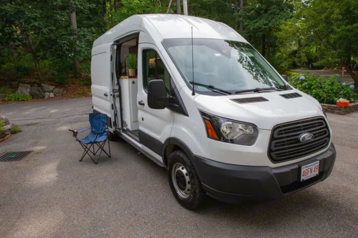 White Ford Transit campervan RV rental parked with side door open, revealing interior. A blue folding chair sits nearby.. Ford Transit 2018