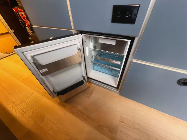 An RV rental interior features an open compact fridge with shelves & blue drawer, a nearby electrical outlet, and visible power wiring.. Mercedes Sprinter 2024