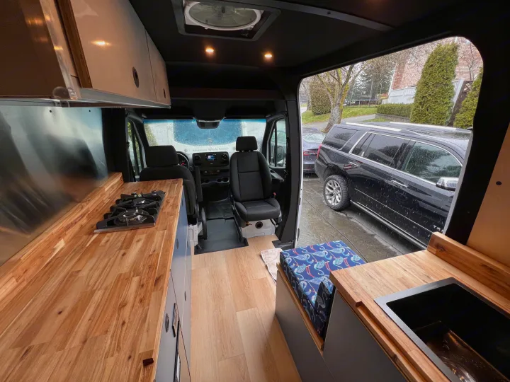 Interior view of a modern RV rental showing a kitchen with wood counters, stove, sink, and a patterned bench.. Mercedes Sprinter 2024