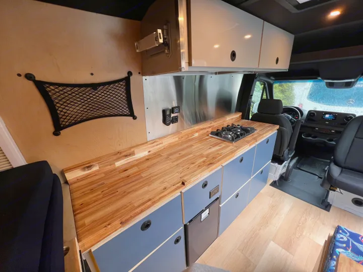 Custom RV rental interior featuring a wood kitchen counter, gas stove, and blue cabinets for mobile living comfort.. Mercedes Sprinter 2024