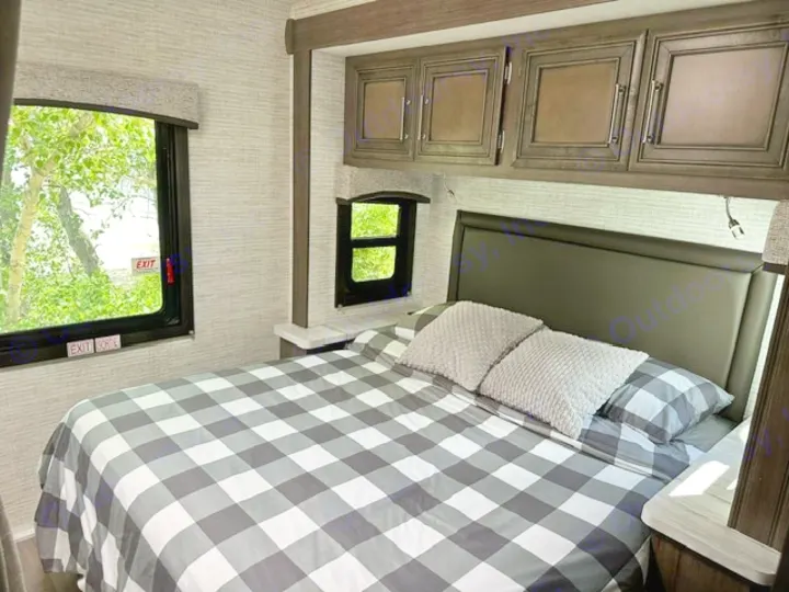 Cozy RV rental bedroom featuring a plaid bed, overhead cabinets, and windows showing green trees outside.. Entegra Coach Odyssey SE 22C 2024