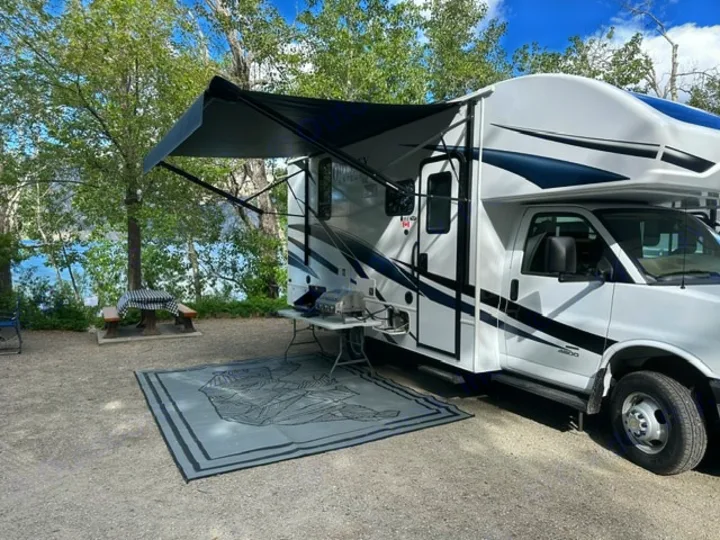 This RV rental is parked at a scenic lakeside campsite, complete with an awning, outdoor mat, grill, and picnic table.. Entegra Coach Odyssey SE 22C 2024