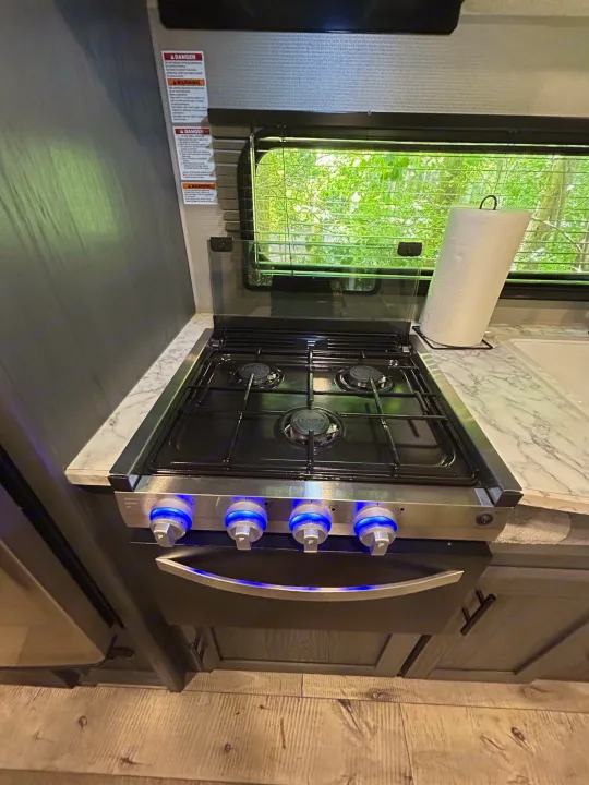 RV rental kitchen: gas stove with blue-lit controls, marble countertop, paper towels, and window view.. Dutchmen Aspen Trail LE 2022