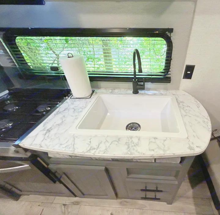 Compact RV rental kitchen interior: white sink, black faucet, marble counter, stove, and window with blinds.. Dutchmen Aspen Trail LE 2022