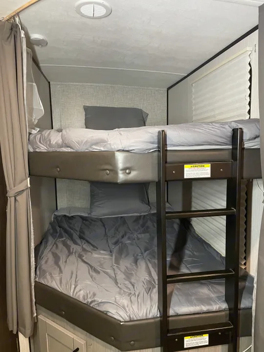 Inside an RV rental: two neatly made bunk beds with gray bedding, a safety ladder, and privacy curtains.. Heartland RVs Mallard 2021