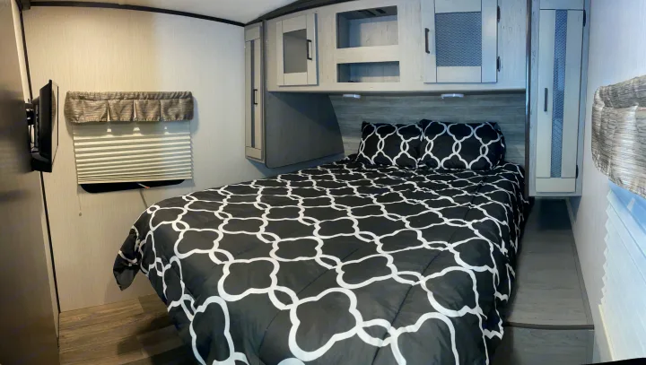 RV rental bedroom with a black & white patterned bed, overhead cabinets, wall-mounted TV, & side storage.. Heartland RVs Mallard 2021