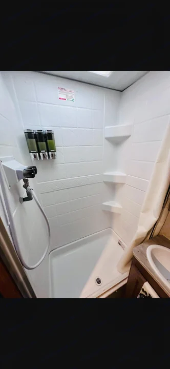 Clean, compact RV rental shower with wall-mounted soap dispenser, corner shelves, and an attached shower head.. No Boundaries 19.8 2021