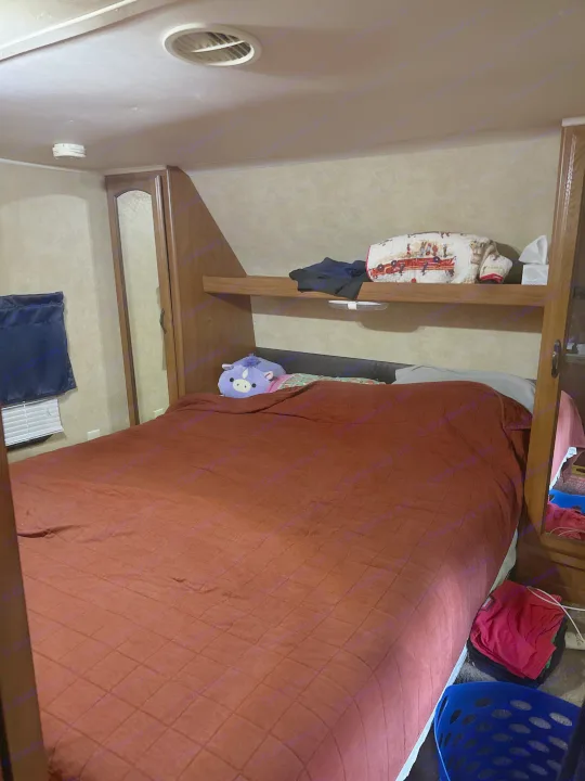 Inside an RV rental bedroom: a cozy bed with red comforter, purple plushie, built-in storage, and window blinds.. Forest River Wildwood 2012