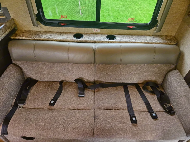 RV rental interior: bench seat with seatbelts, cupholders on the counter, and an emergency exit window overlooking grass.. Thor Freedom Elite 2018