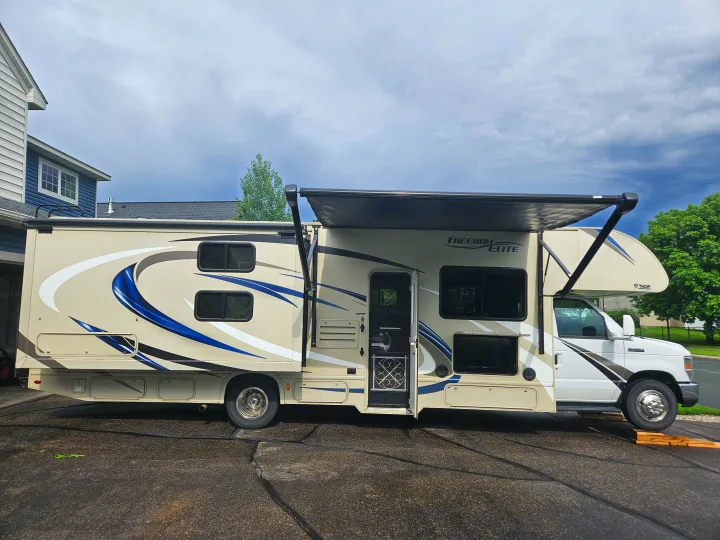 This Class C Thor Freedom Elite RV rental, beige with blue graphics, has its awning extended and door open on a driveway.. Thor Freedom Elite 2018