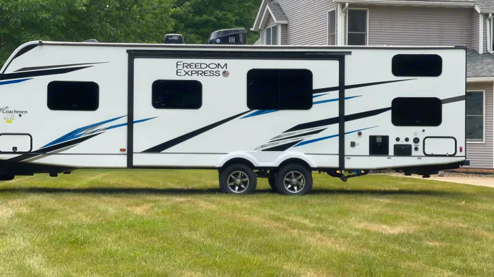 Detailed side view of a white Freedom Express travel trailer RV rental, featuring blue/black graphics & multiple windows, parked on grass.. Coachmen 292BHDS FREEDOM EXPRESS LIBERTY EDITION 2022