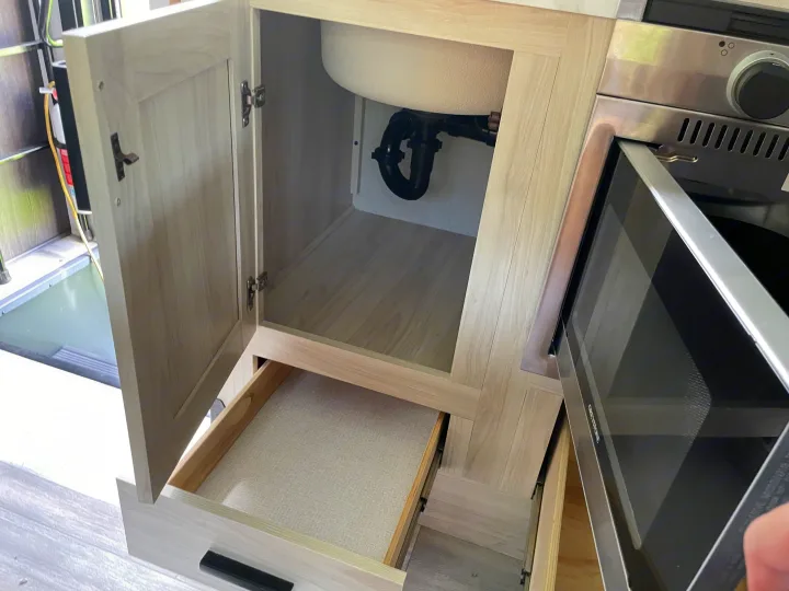 RV rental kitchen: open sink cabinet, drawer with liner, oven, and plumbing. Fire extinguisher visible left.. Forest River Sunseeker LE 2023