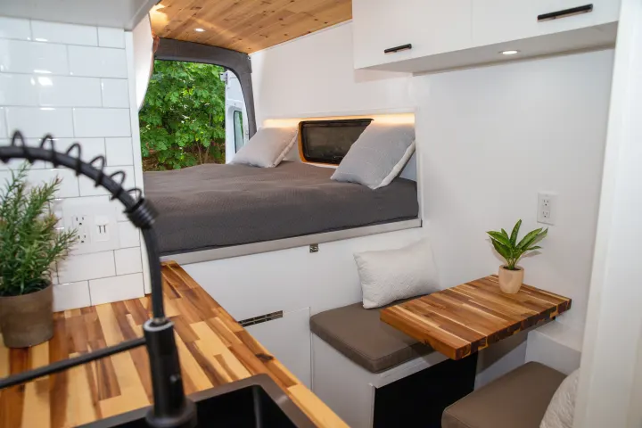 Modern RV rental interior with a raised bed, full kitchen, and dinette. Features a sleek design with wood accents and white walls.. Ford Transit 2018