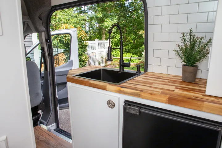 This RV rental's van conversion kitchen has a sleek black sink, wood countertop, and a view to nature.. Ford Transit 2018