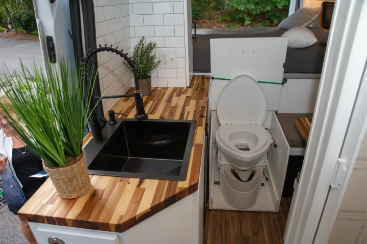 Compact RV rental interior with a modern kitchen, black sink, and a functional toilet setup. Features bed & wood accents.. Ford Transit 2018