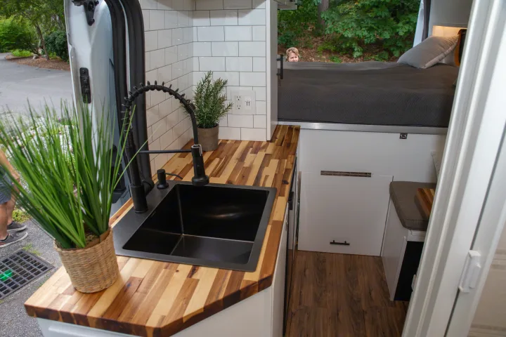 Bright rv rental van interior: a modern kitchen with black sink, wood counters, subway tile, and a cozy bed setup.. Ford Transit 2018