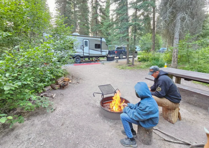 A family enjoys a warm campfire at their RV rental campsite, with a travel trailer and truck visible in the forest setting.. Jayco Jay Flight 232 RBW with Rocky Mountain and Baja upgrades 2020