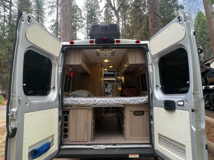 Rear view of an RV rental camper van, doors open to reveal a comfortable bed, light wood cabinetry, and integrated controls.. Thor Motor Coach Sequence 2022