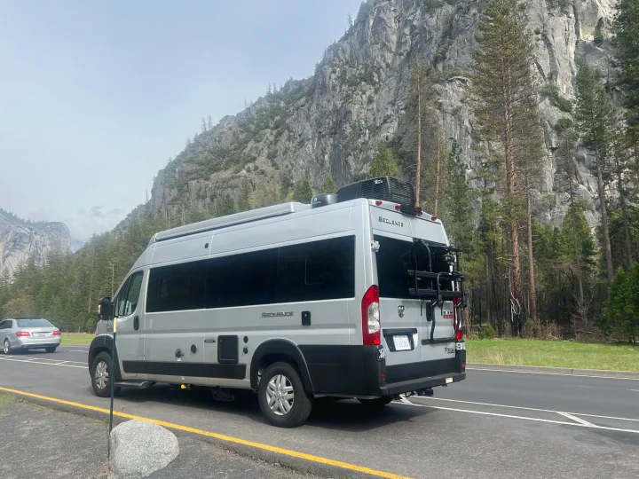 A grey Thor Sequence camper van (RV rental) with a bike rack is parked on a scenic road with large mountain cliffs and trees.. Thor Motor Coach Sequence 2022