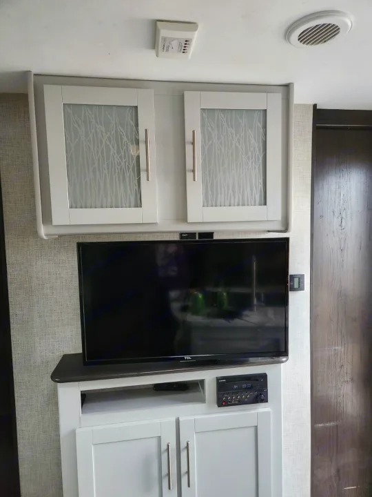 An RV rental interior featuring an entertainment setup with a TV, media console, and overhead cabinets. Ceiling vent and detector visible.. Keystone RV Bullet Ultra Lite 2020