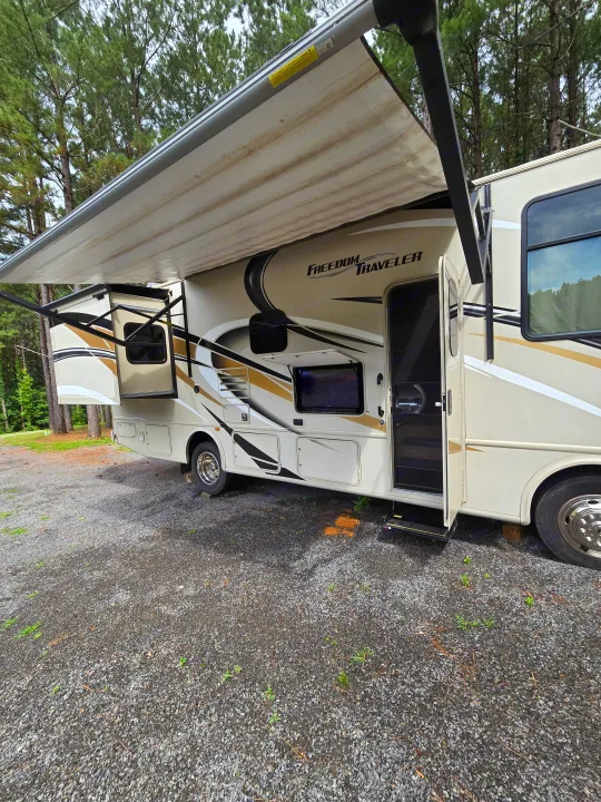 Modern rv rental parked on gravel at a forest campsite, with its awning extended and a slide-out open.. Thor Freedom Traveler 2020