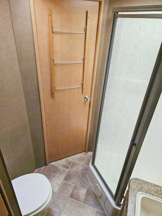 Compact RV rental bathroom featuring a white toilet, tiled floor, shower stall with a textured glass door, and a door with a towel rack.. Thor Chateau 2016