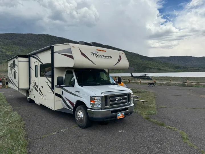 Scenic rv rental adventure: Class C RV parked lakeside with mountains & a helicopter nearby under cloudy skies.. Coachmen Freelancer 2019