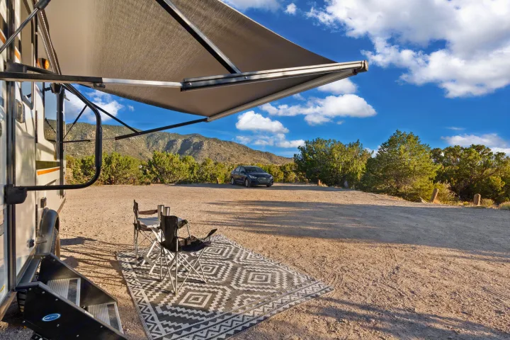 RV rental parked with awning extended, outdoor rug, chairs, and table set up. Car and mountains in background.. Rockwood G19QB 2021