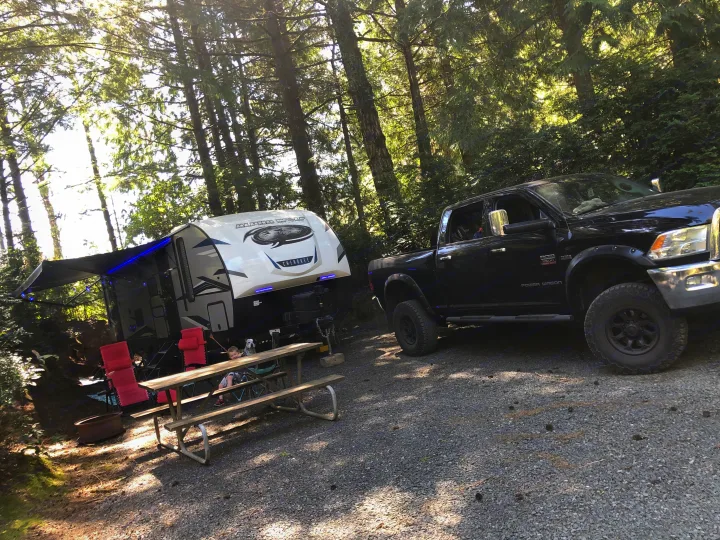 Perfect RV rental setup! Black truck & travel trailer at a sunny, treed campsite with a picnic table, fire pit, and family.. Forest River Cherokee Alpha Wolf 2021