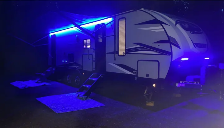 A modern RV rental glows bright blue at night from its awning lights & exterior accents, set up for a campsite stay.. Forest River Cherokee Alpha Wolf 2021