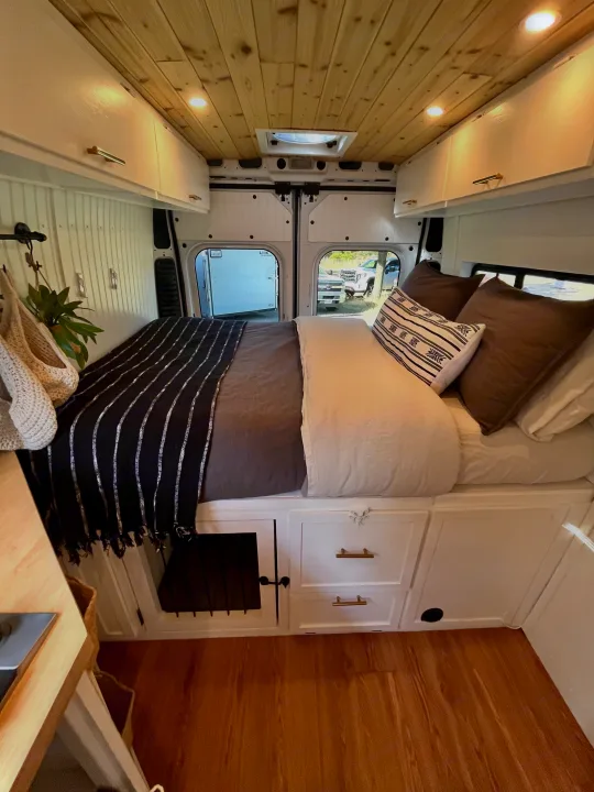 Cozy RV rental interior with a bed, wood ceiling, white cabinets, and outdoor views for comfortable travel.. Dodge B Van 2015