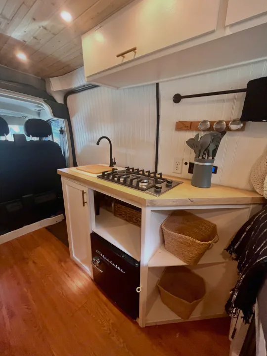 Inside an RV rental: a compact kitchen area with wood accents, featuring a stove, sink, and built-in storage.. Dodge B Van 2015