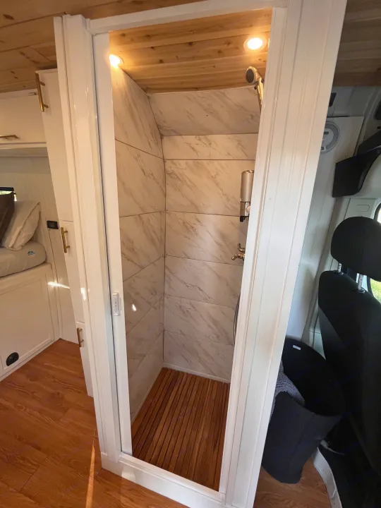Compact, modern shower inside an RV rental, featuring marble-look walls, wood floor, and recessed lighting, perfect for van life.. Dodge B Van 2015