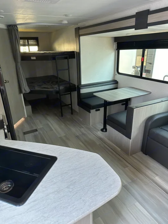 This RV rental shows bunk beds, a dinette with seating, and a kitchen counter. Bright interior with wood-look flooring.. Keystone RV SG261BHC 2024