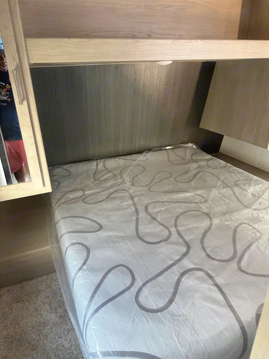 RV rental bed area: A new mattress with plastic wrap, sleek wood-grain interior, and overhead storage for travel.. Keystone RV SG261BHC 2024
