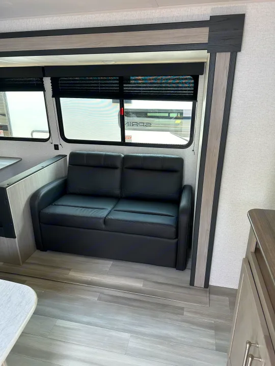 Comfortable RV rental living space featuring a black loveseat, modern light wood trim, grey wood-look flooring, and windows.. Keystone RV SG261BHC 2024