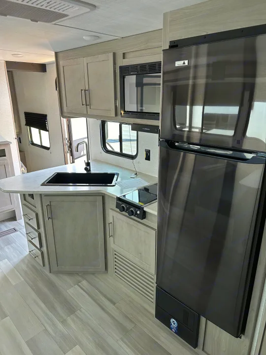 Bright RV rental kitchen: light wood-tone cabinets, dark stainless appliances (fridge, microwave, stovetop), sink, light flooring.. Keystone RV SG261BHC 2024