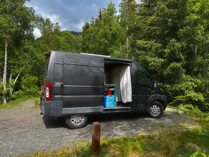 Dark gray rv rental van parked on gravel, side door open to reveal interior, surrounded by lush green trees and distant mountains.. Dodge Ram 1500 2017